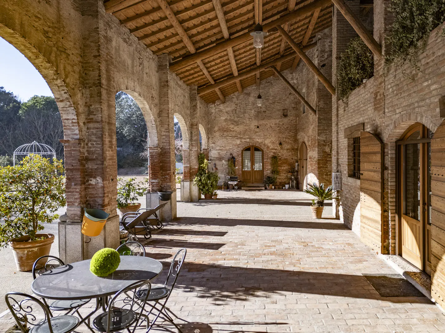 Rustic brick patio with arched columns, wooden ceiling beams, outdoor seating, and potted plants in a sunlit courtyard.