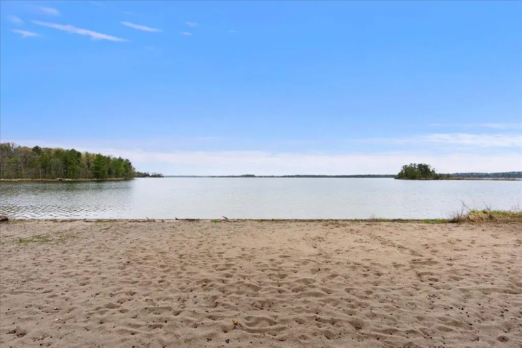 Sandy beach leading to a calm lake under a blue sky. Trees line the shore in the distance.