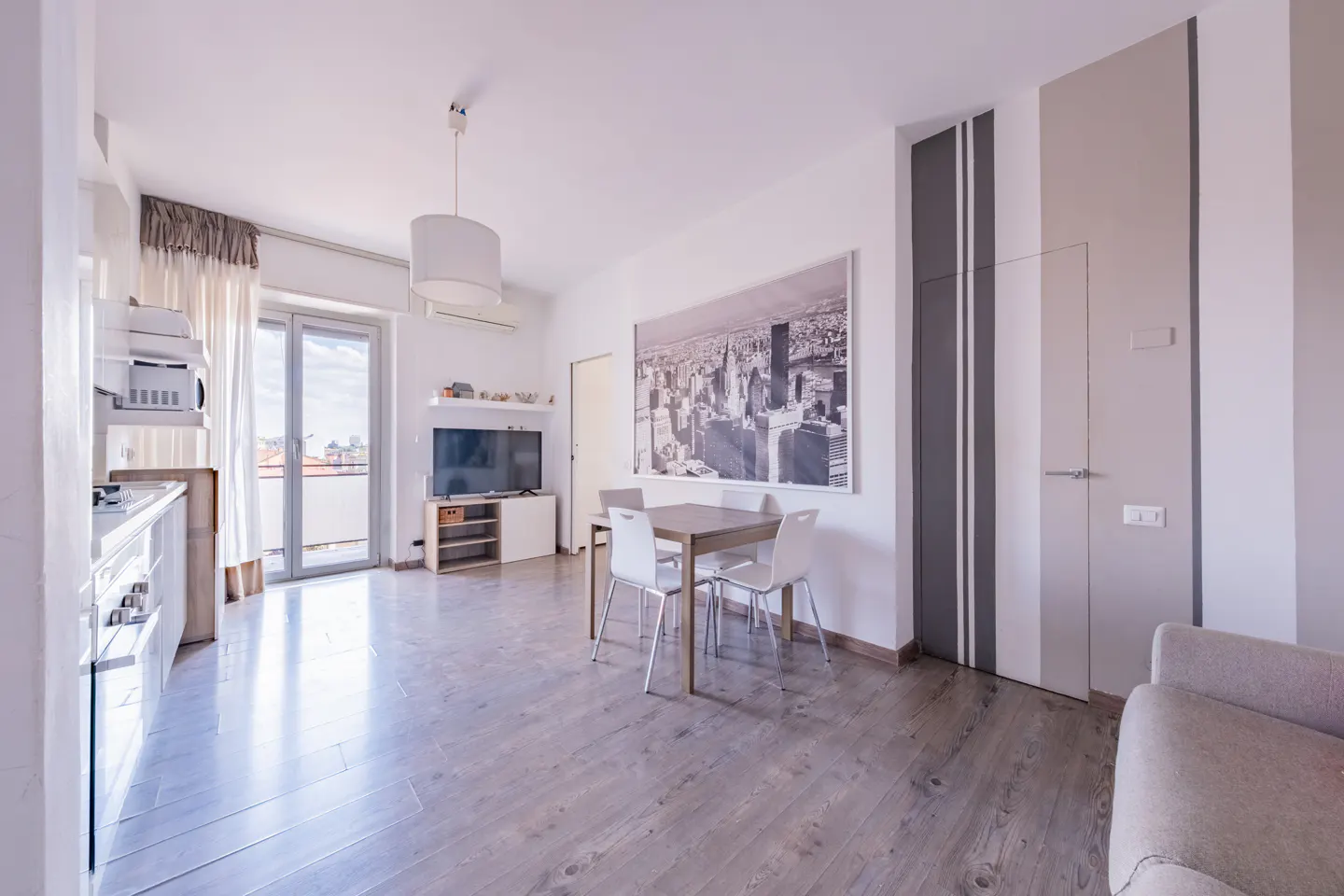 Bright, modern apartment interior with wood floors, white walls, and a dining table. A black and white cityscape print hangs on the wall.