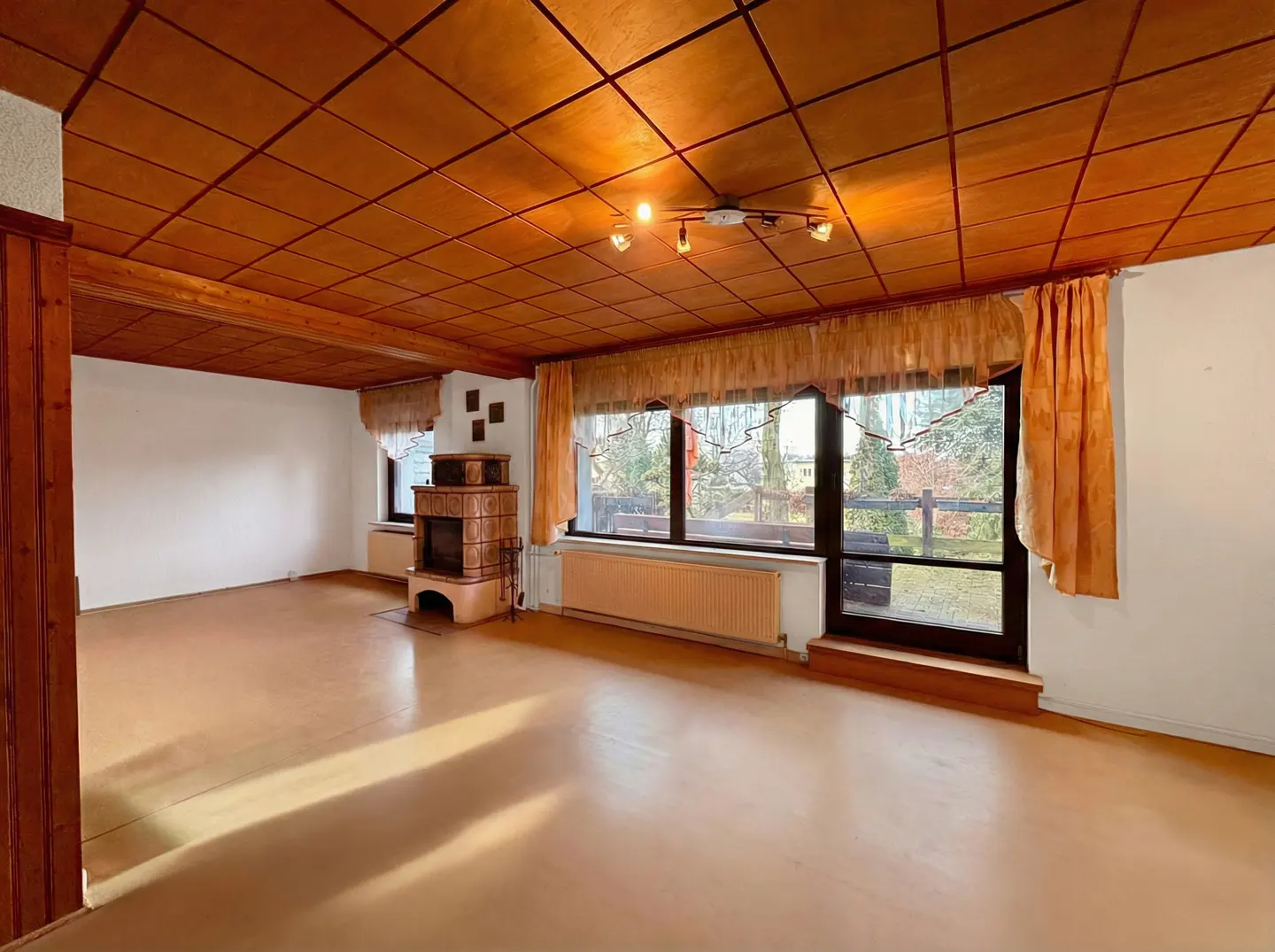 A spacious living room with a wood-paneled ceiling, a tiled fireplace, and large windows with orange curtains.