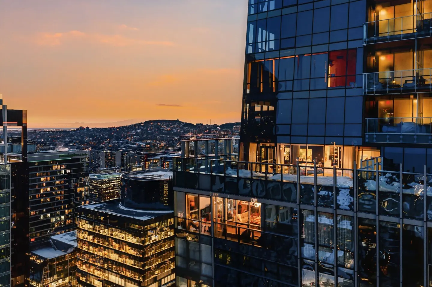 Cityscape view at sunset, featuring modern glass skyscrapers with illuminated interiors and a distant mountain range.