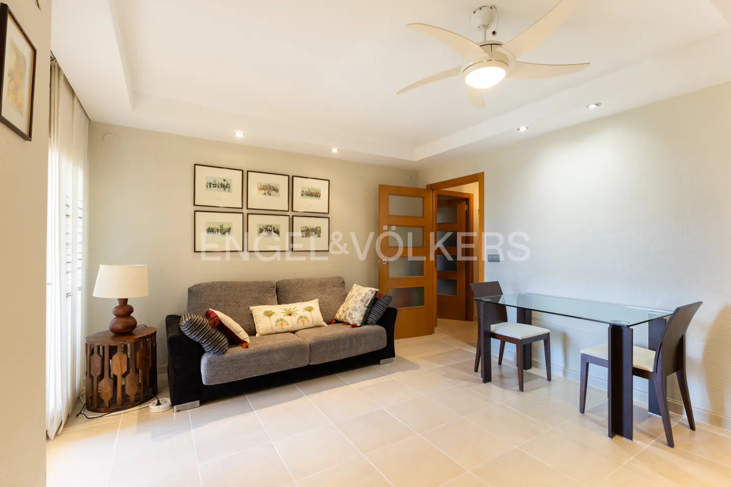Living room with a gray sofa, glass table, and wooden chairs. A ceiling fan is visible. The walls are light beige.