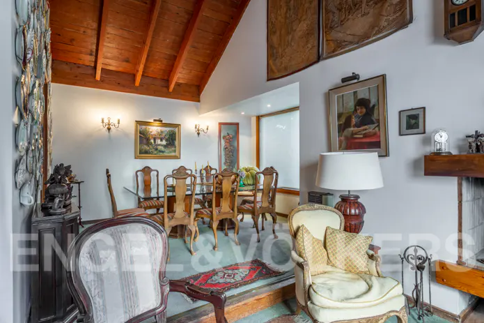 A living room with a dining area, featuring wooden furniture, paintings, and a high, wooden ceiling.