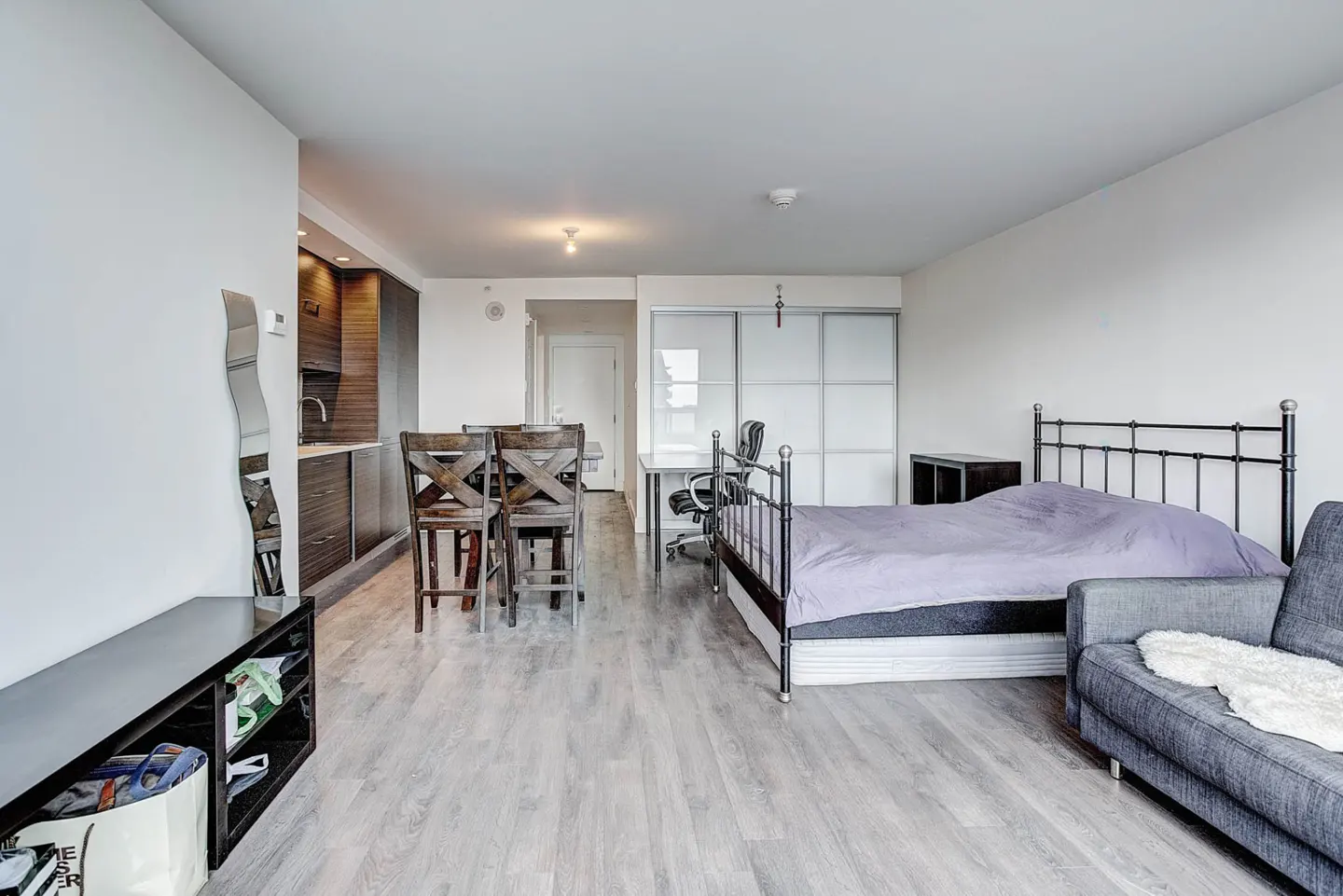 Studio apartment with a bed, grey sofa, dining table, and kitchenette with dark wood cabinets on light wood flooring.