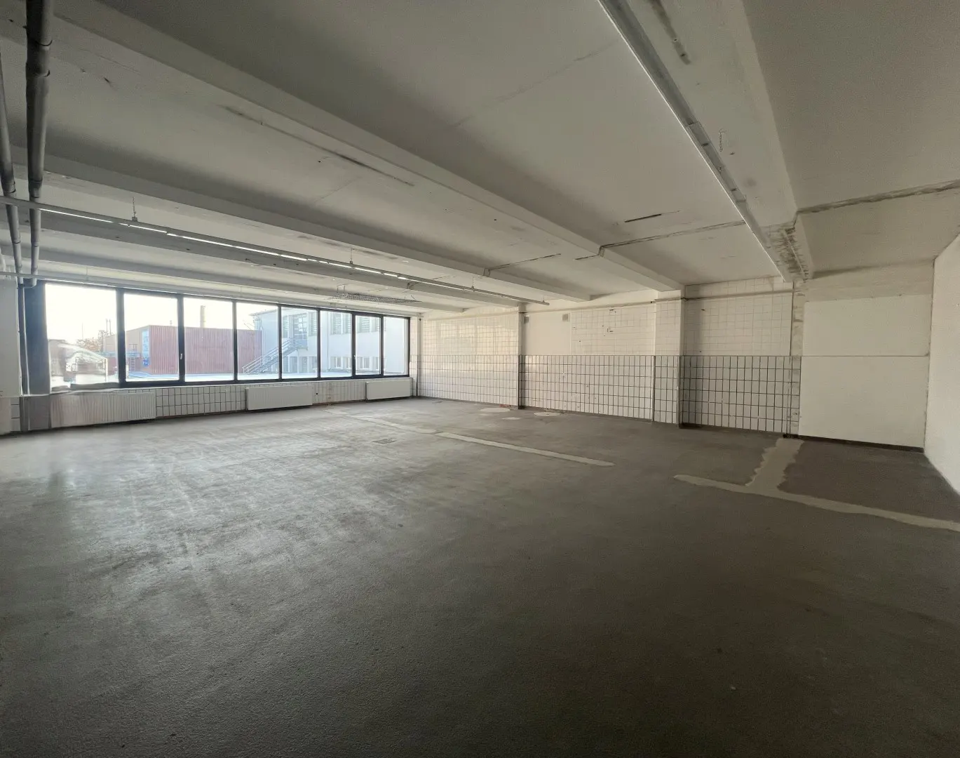 An empty, industrial-style commercial space with concrete floors, white tiled walls, and large windows overlooking a street.