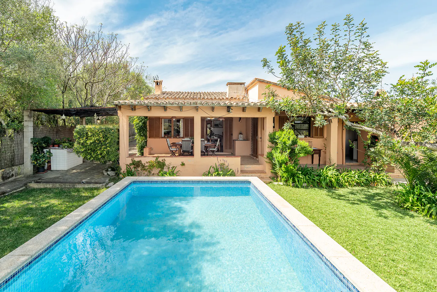 A tan house with a tiled roof overlooks a blue swimming pool and green lawn under a partly cloudy sky.