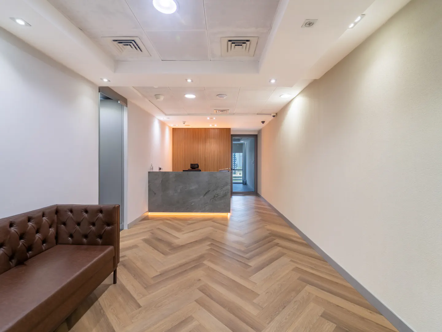 Office lobby with a brown leather sofa, herringbone wood floors, and a gray reception desk with wood paneling behind it.