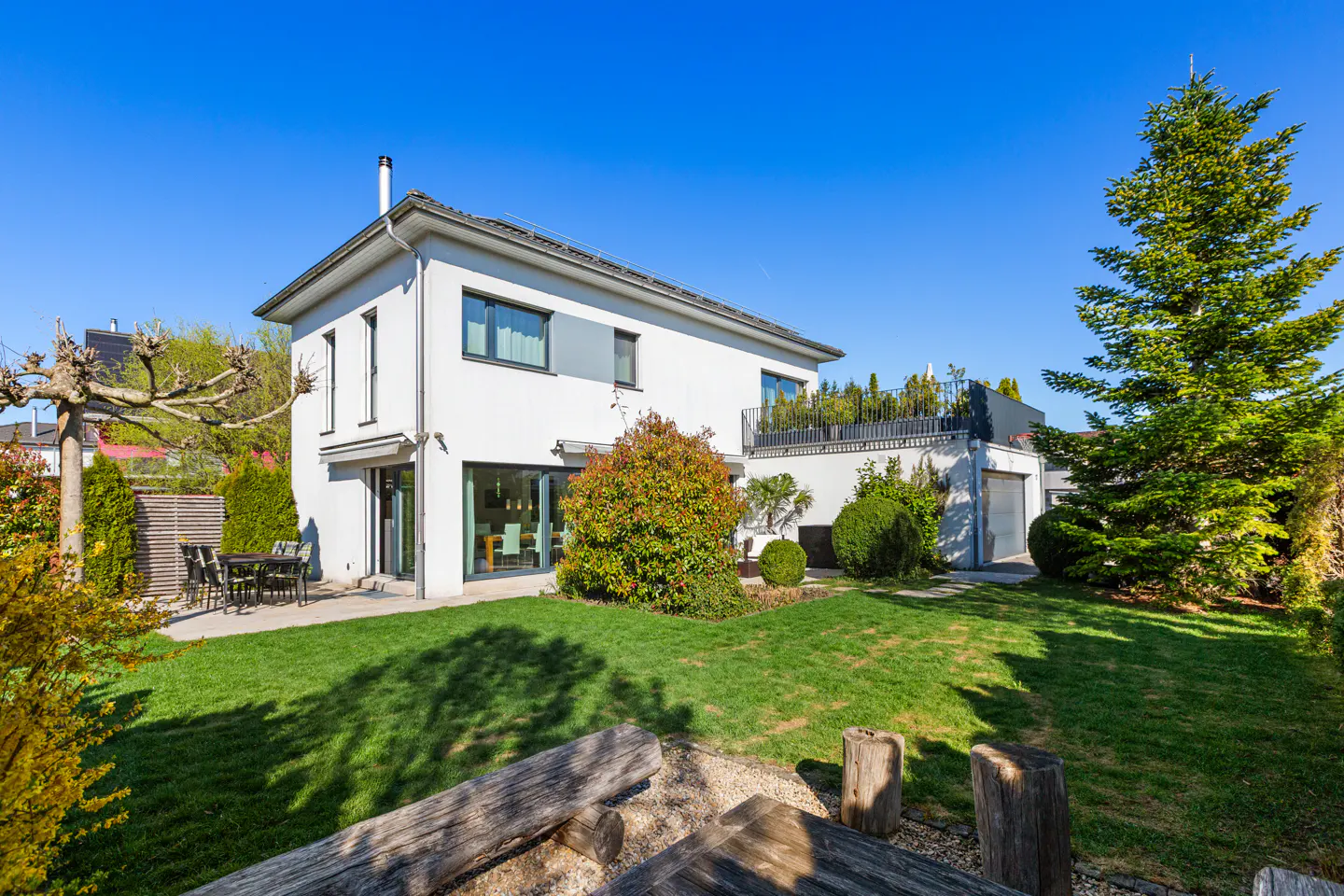 Two-story white house with a green lawn, trees, and a blue sky. Outdoor furniture is visible on the patio.