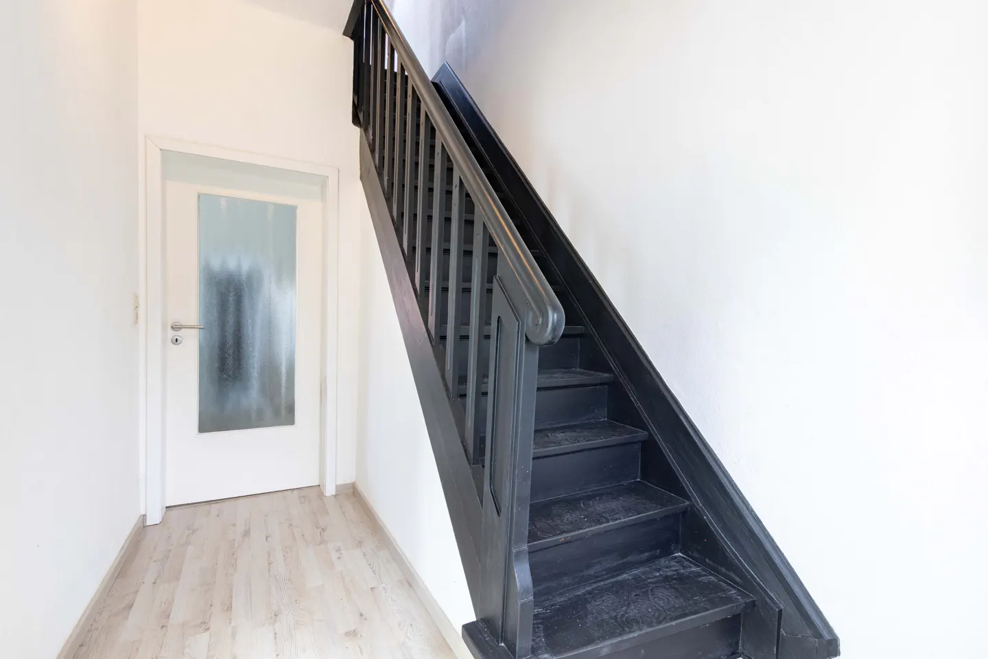 Interior view of a hallway with a black staircase and a white door with a frosted glass panel. The walls are white, and the floor is light wood.