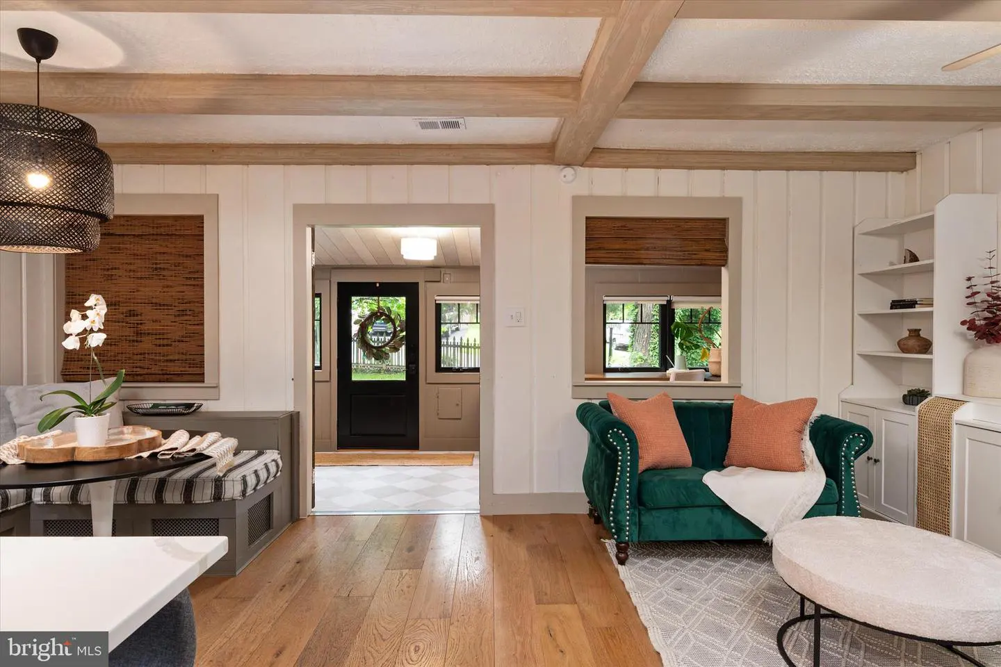 Living room with wood floors, exposed beams, and a green velvet loveseat with orange pillows. A black front door is visible in the background.