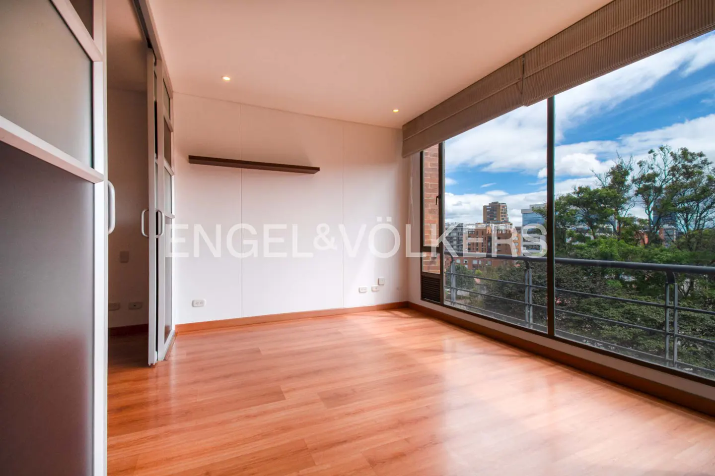 Empty room with wood floors, white walls, and a large window with a city view. Engel & Völkers logo on the wall.