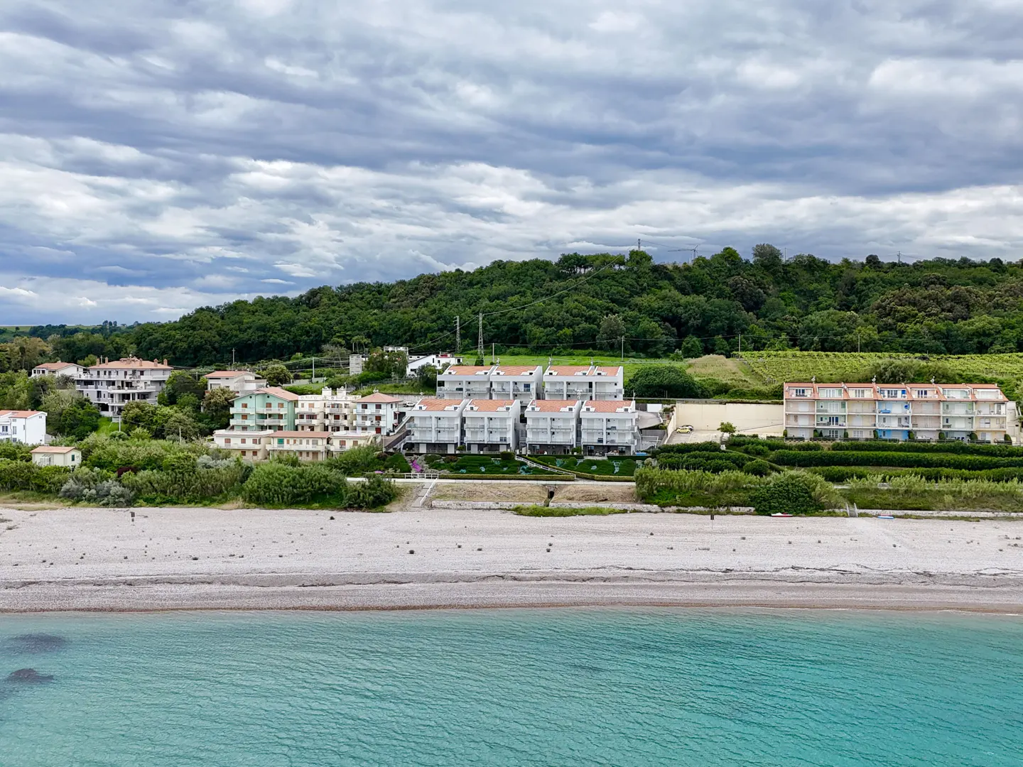 Coastal view of multi-story condos with red roofs, a pebble beach, and turquoise water under a cloudy sky.