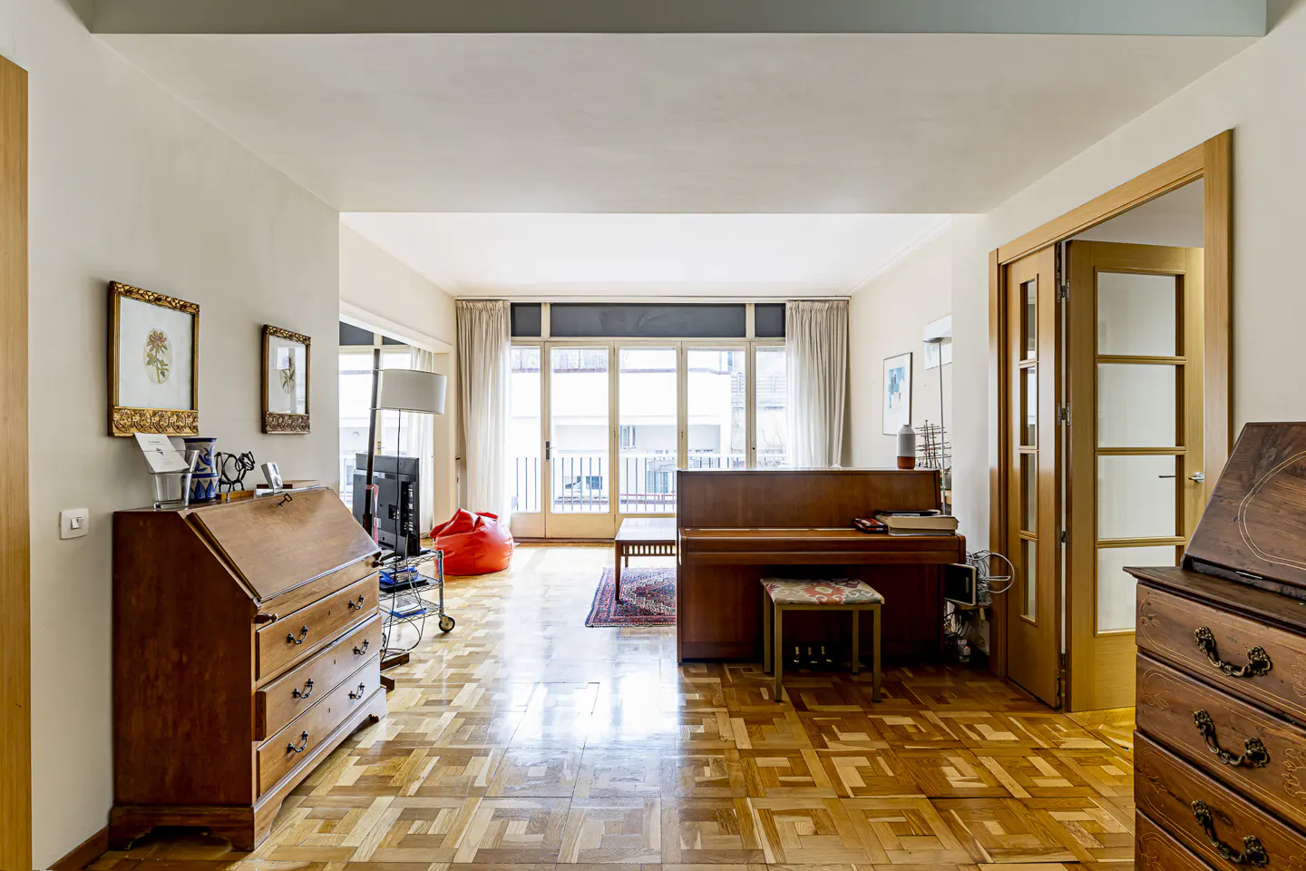 Living room with parquet floors, a wooden piano, two antique desks, and a large window leading to a balcony.