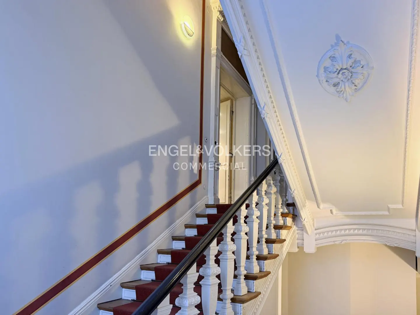 Interior view of a staircase with red carpet, white balusters, and a black handrail. Ornate white molding adorns the walls and ceiling.