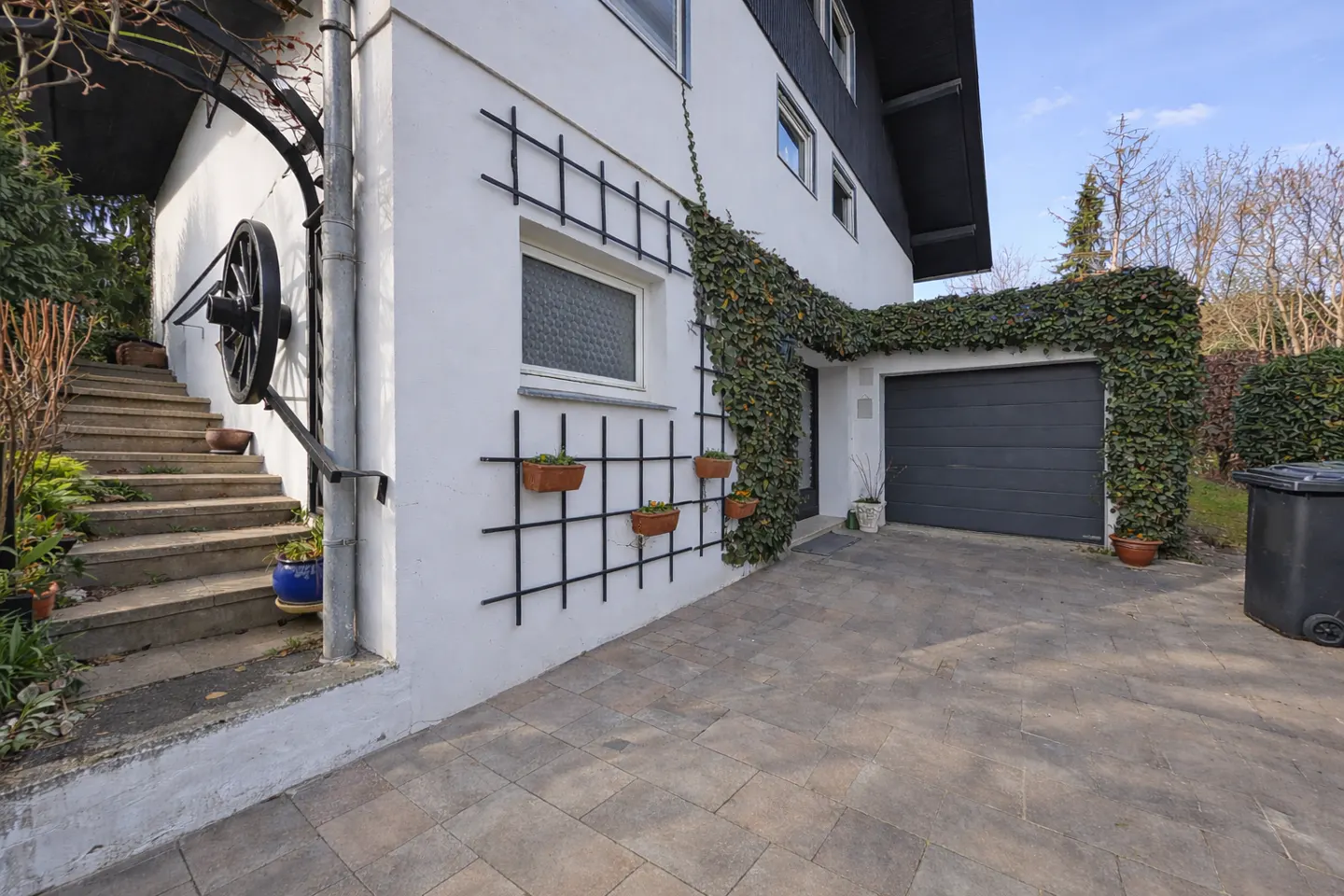 White house exterior with a paved driveway, garage, stone stairs, and ivy-covered walls with decorative wall planters.