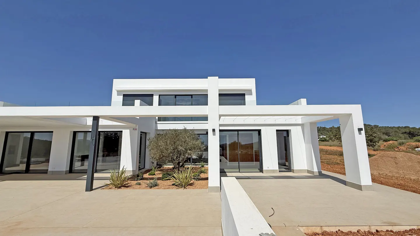 Modern white two-story house with a flat roof, large windows, and a pergola over the patio. Blue sky background.