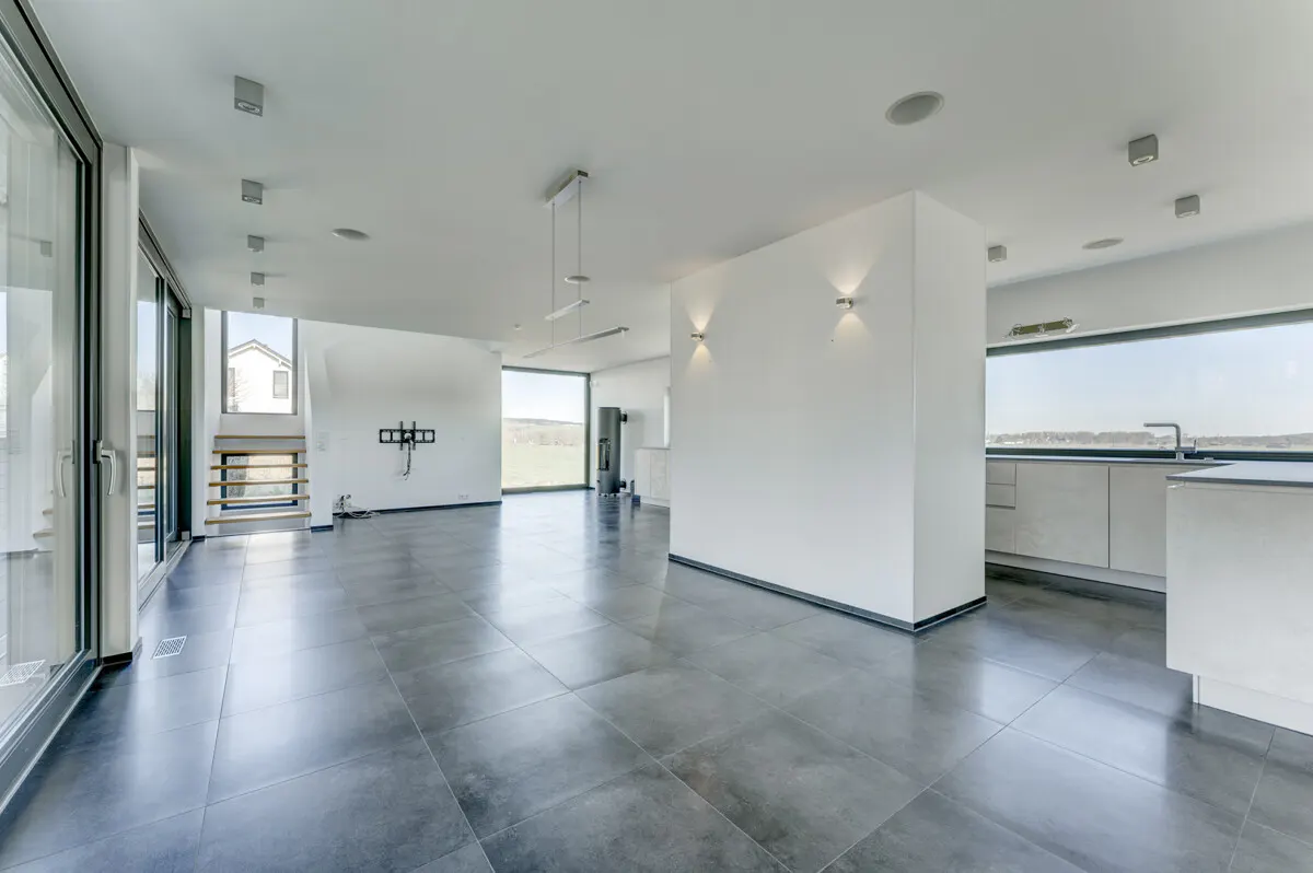 Bright, modern open-plan living space with gray tile floor, white walls, and large windows. Kitchen area visible in the background.