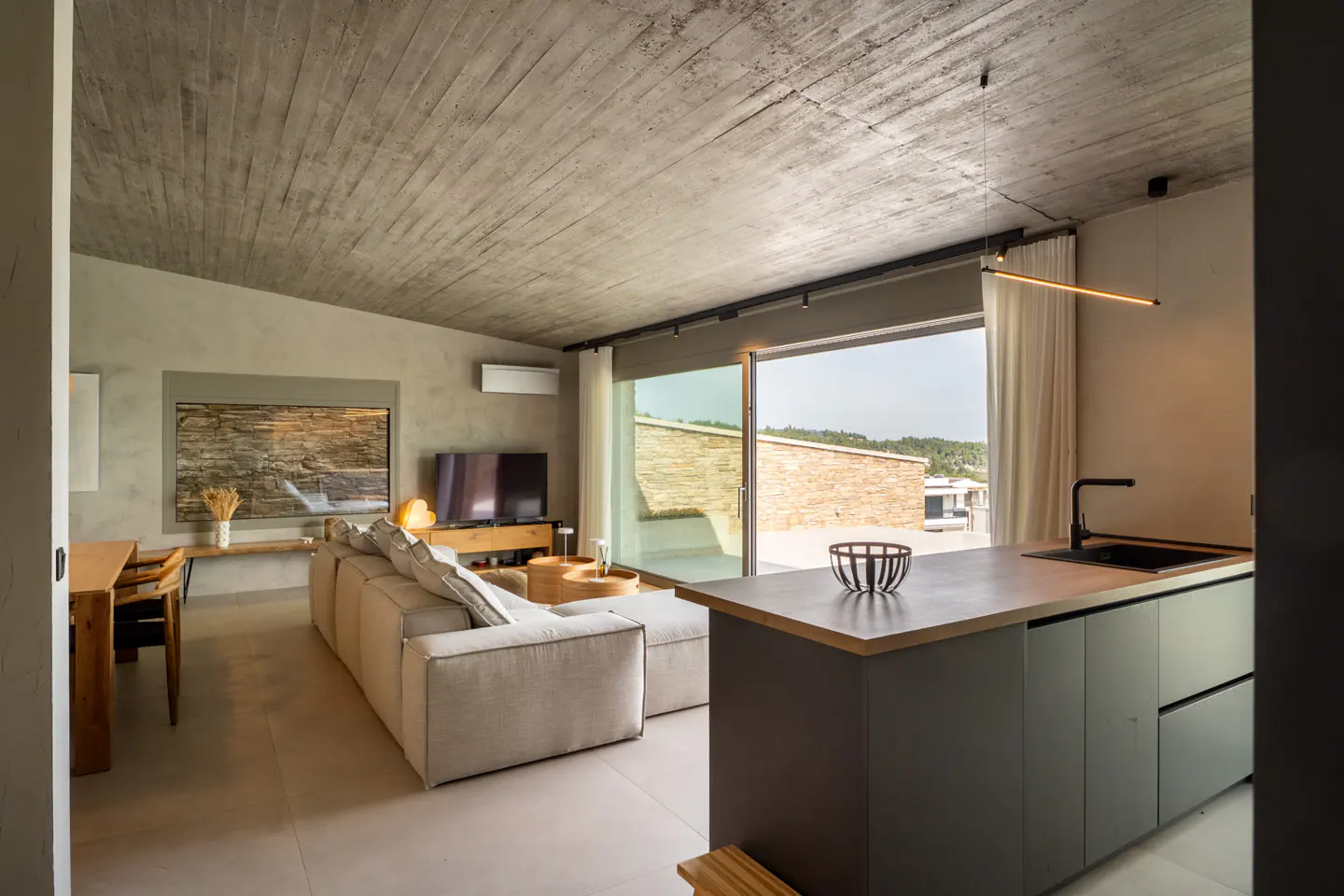 Modern living room with concrete ceiling, gray kitchen island, beige sofa, TV, and sliding glass doors to a patio.