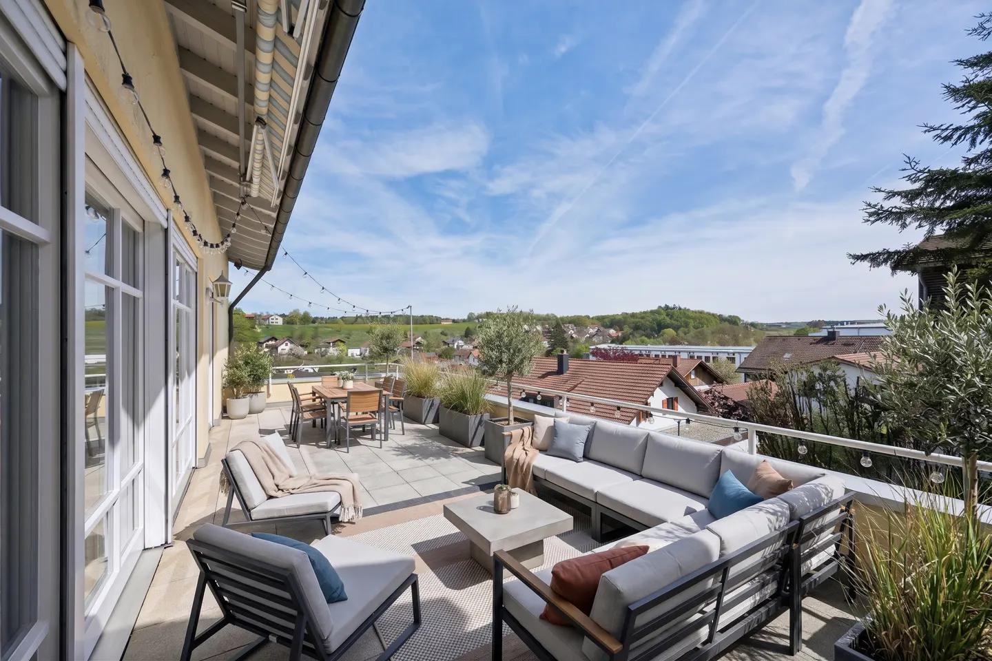 A rooftop patio with gray furniture, a dining table, and potted plants overlooks a town with a blue sky.