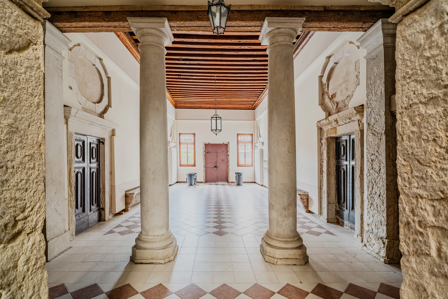 Hallway with stone columns, wood ceiling, and diamond-patterned tile floor. A red door is centered at the end of the hall.