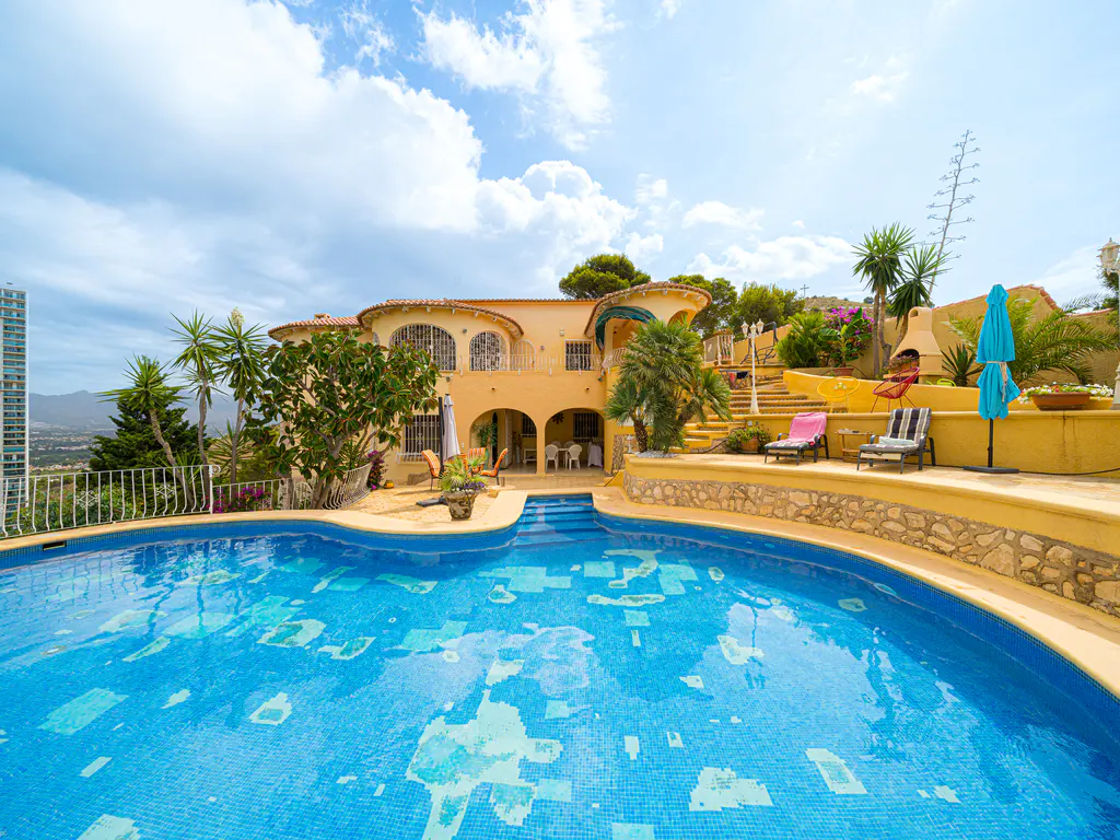 A sunny Mediterranean villa with a blue swimming pool, stone patio, lounge chairs, and lush palm trees under a cloudy sky.