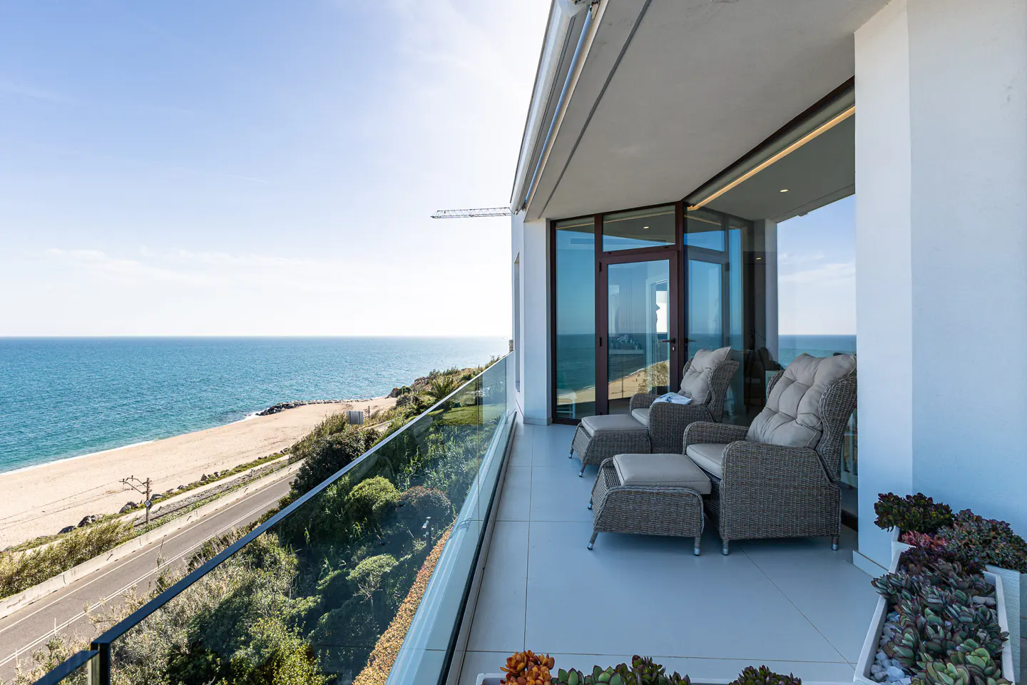 Balcony with wicker chairs and ocean view. Glass railing overlooks beach, road, and blue sea under a clear sky. Succulents in pots add greenery.