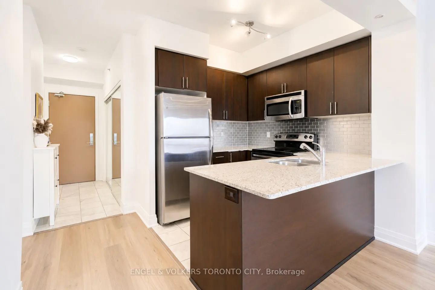 A bright kitchen with dark wood cabinets, stainless steel appliances, and a granite countertop island. A hallway leads to a brown door.