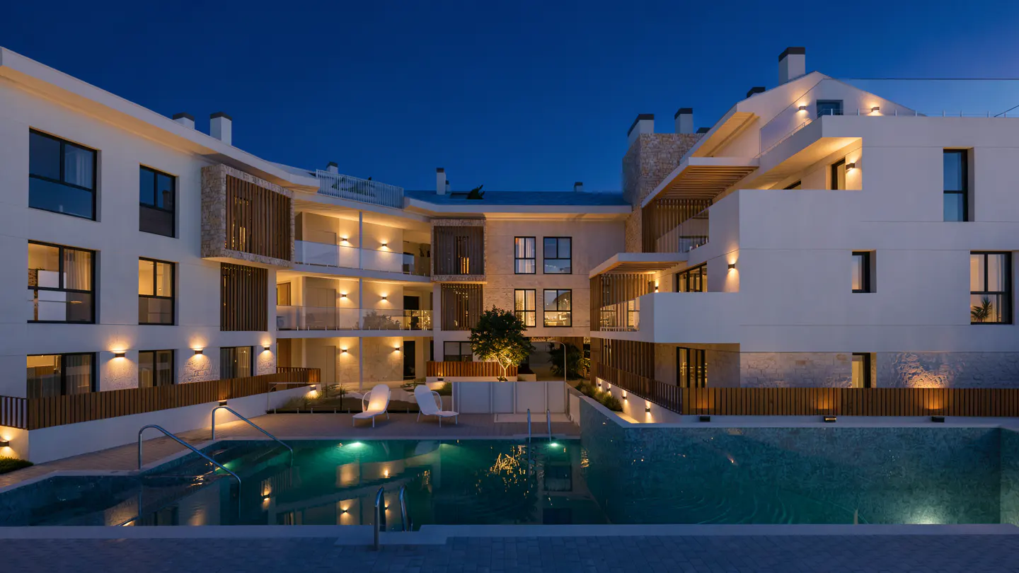 Night view of a modern white apartment complex with a pool, lit by warm lights. Two white chairs sit near the pool.