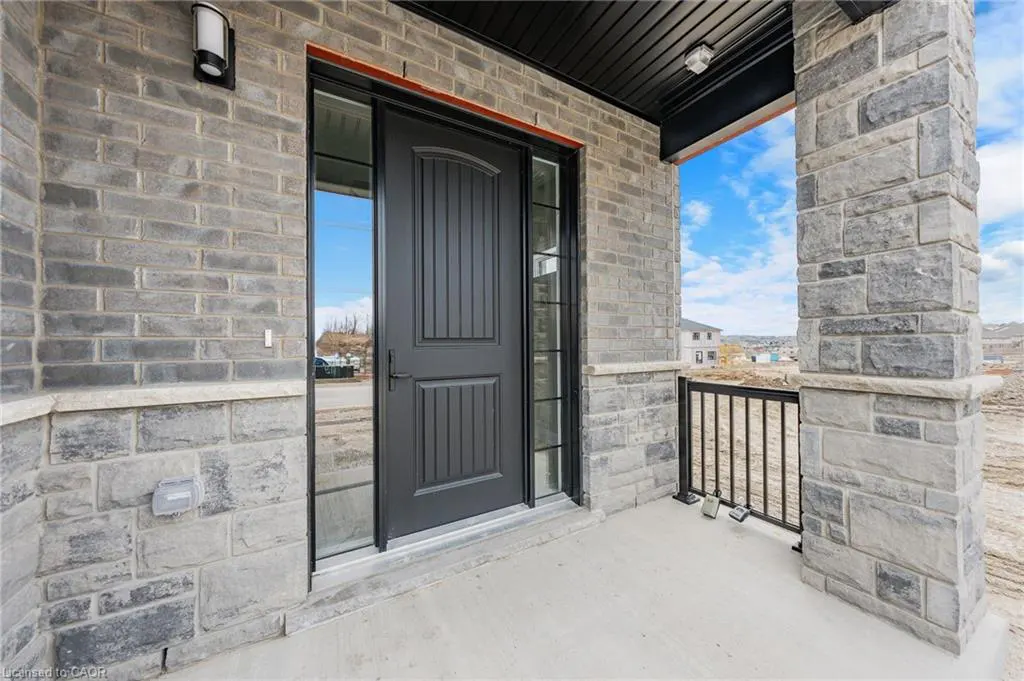 Front entrance of a grey brick house with a black door and stone pillars on a concrete porch.