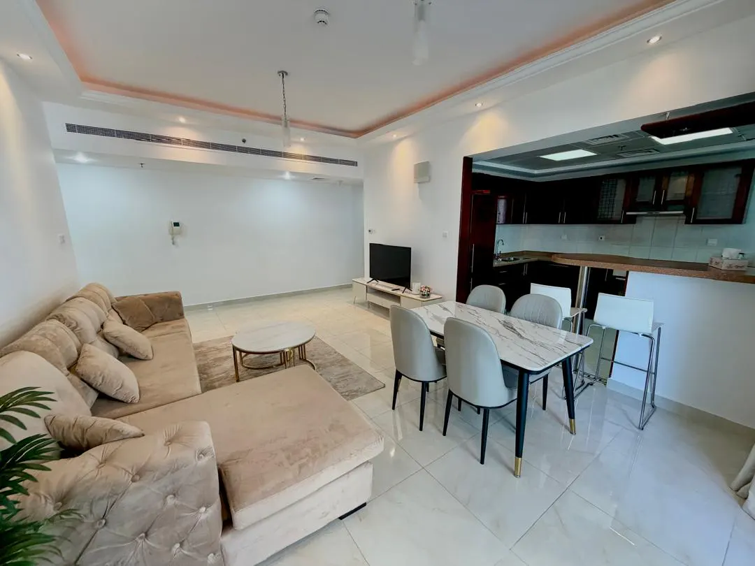 Open-plan living area with a beige tufted sectional sofa, marble dining table, and dark wood kitchen cabinets.
