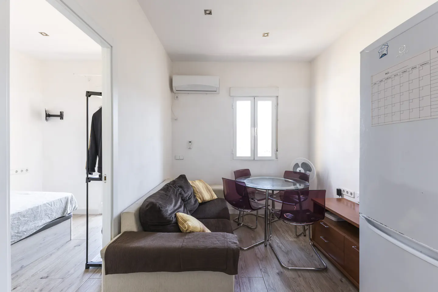 Bright studio apartment with a brown sofa, glass table with purple chairs, and a white refrigerator with a calendar. A bedroom is visible through a doorway.