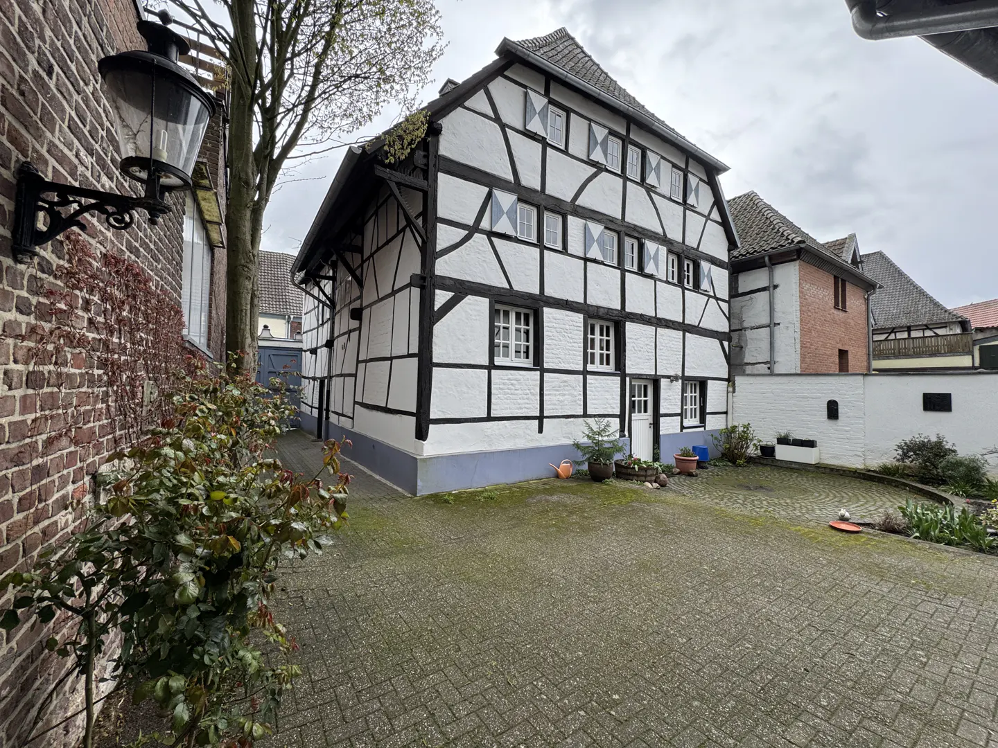 A traditional half-timbered house with white walls and black beams stands beside a paved courtyard and brick wall.