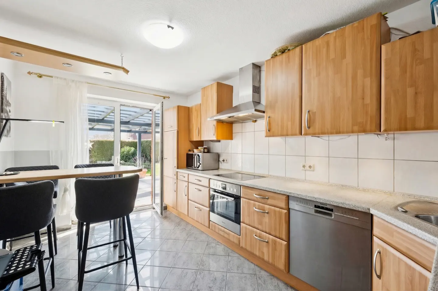 A bright kitchen with light wood cabinets, white tile backsplash, and stainless steel appliances. A table with black chairs sits near a glass door to the outside.