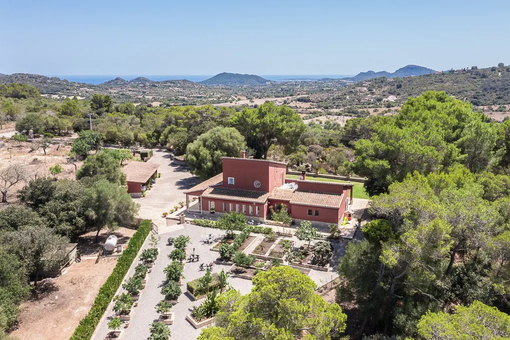 Aerial view of a red-walled house with a tile roof, surrounded by trees and a garden, with mountains and the sea in the background.