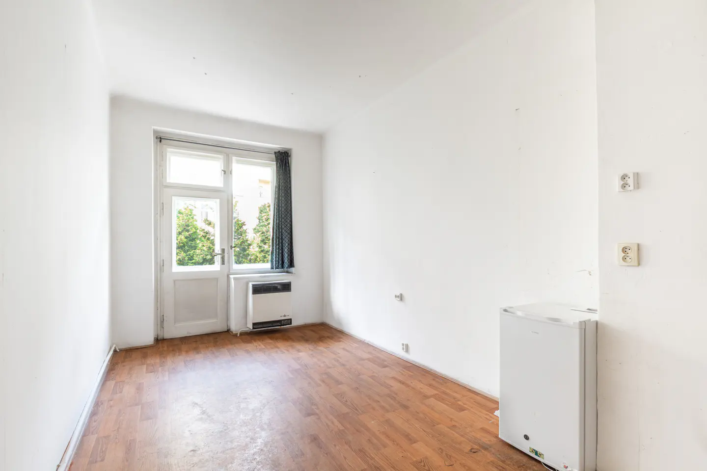 Empty room with white walls, wood-look flooring, a glass door to a balcony, a small heater, and a white mini-fridge.