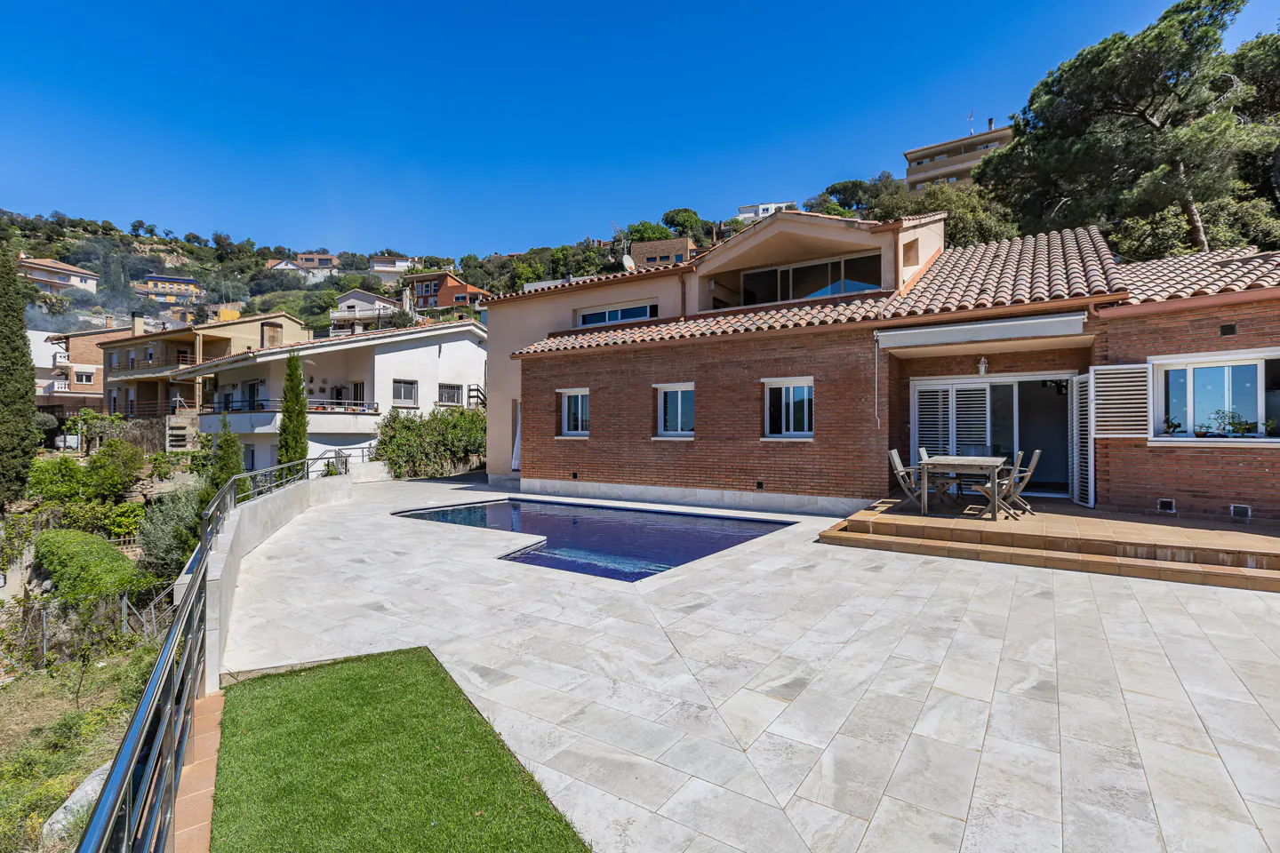 Exterior view of a brick house with a blue pool and patio furniture on a sunny day. Houses are on a hill in the background.