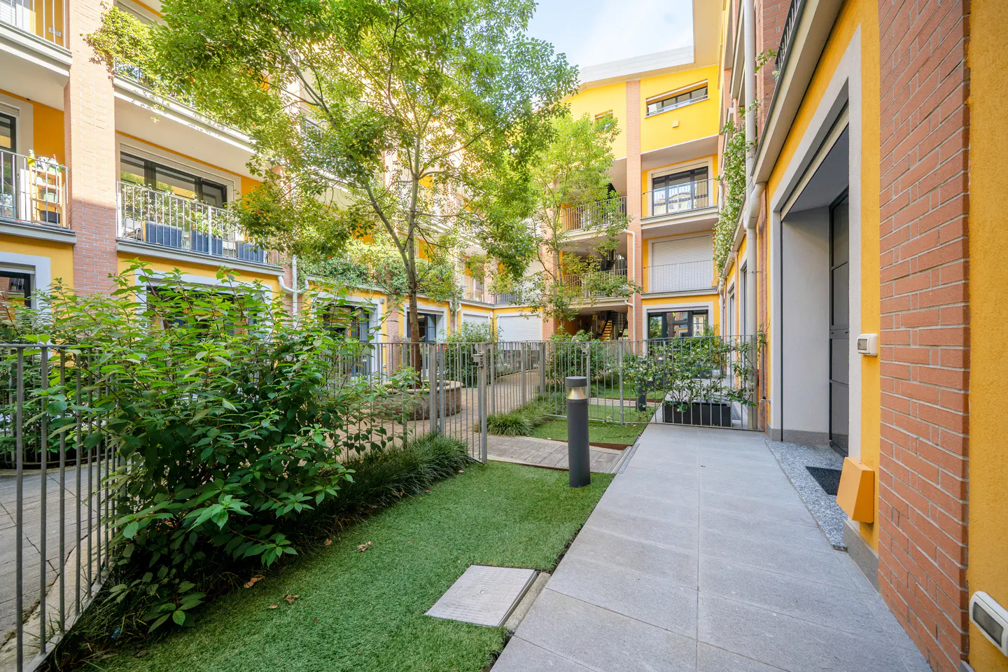 A sunny courtyard with yellow walls, a paved walkway, green lawn, and lush trees surrounded by multi-story apartments.