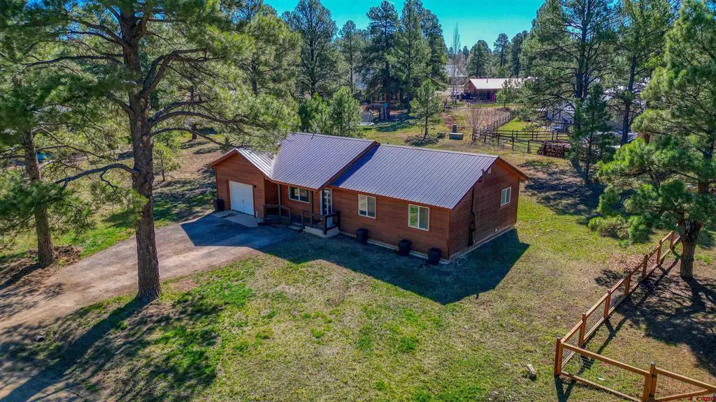 Aerial view of a brown, one-story house with a gray metal roof, surrounded by green trees and grass. A wooden fence is visible in the foreground.