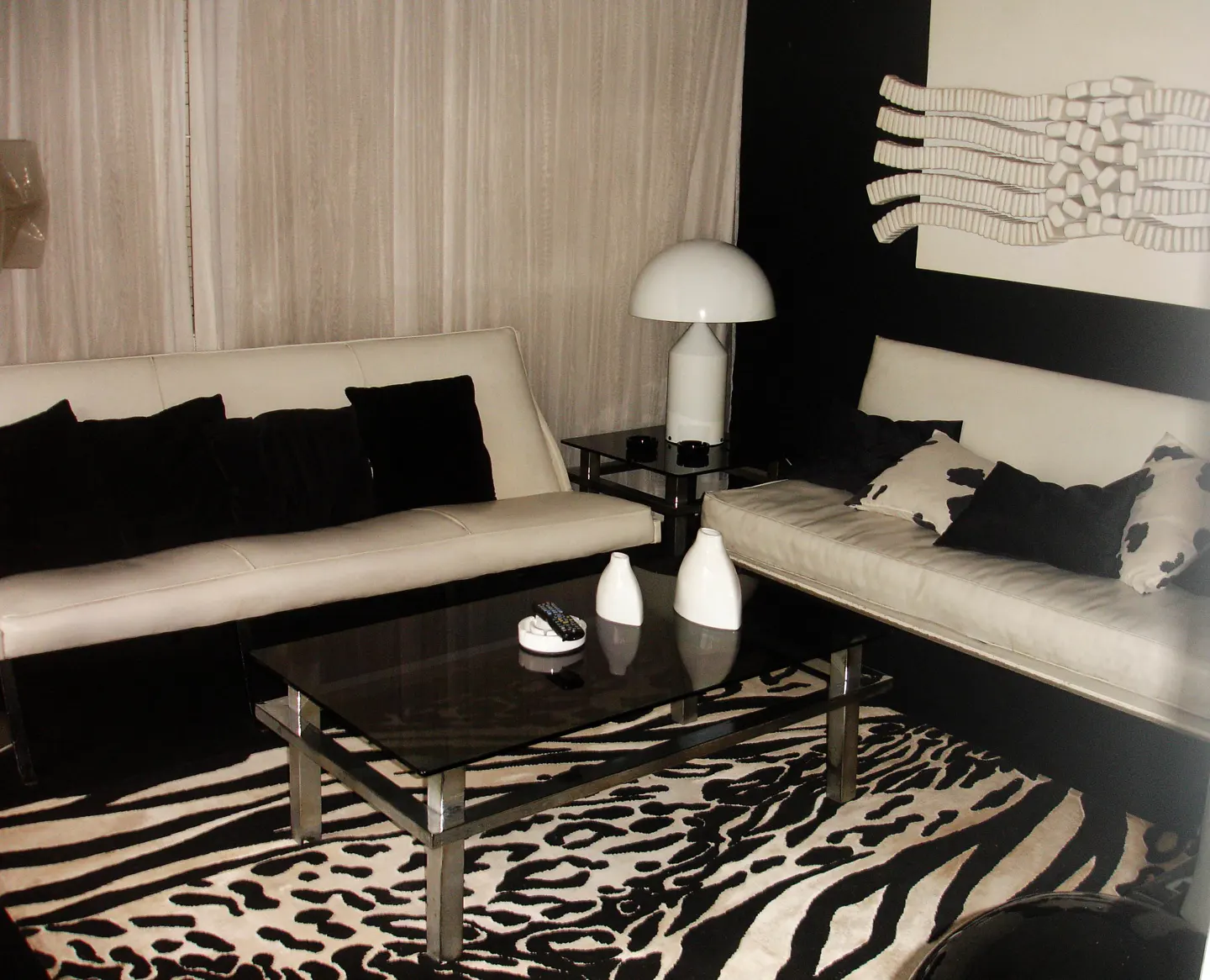 A modern living room with white sofas, black pillows, and a zebra-print rug. A black glass coffee table sits in the center.
