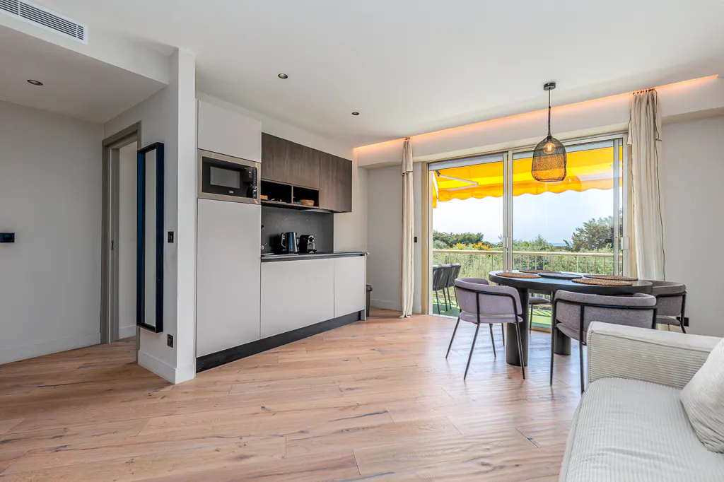 Bright apartment interior with wood floors, a modern kitchen, and a dining area with a view of trees and a yellow awning.