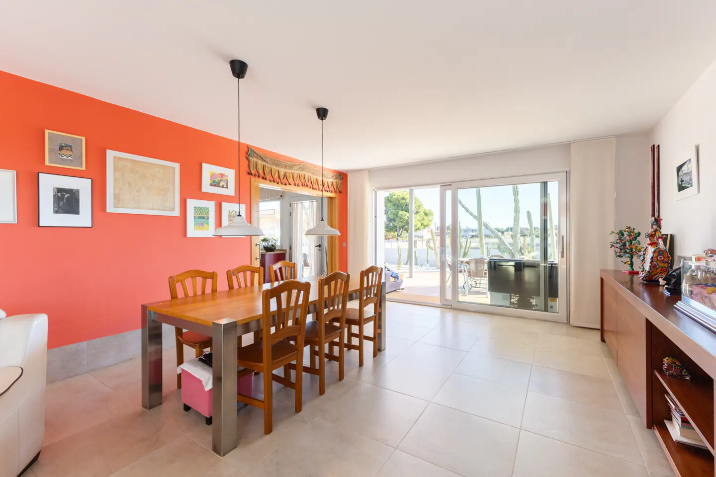 Bright dining room with orange wall, wood table, and chairs. Sliding glass doors lead to a patio with cacti.