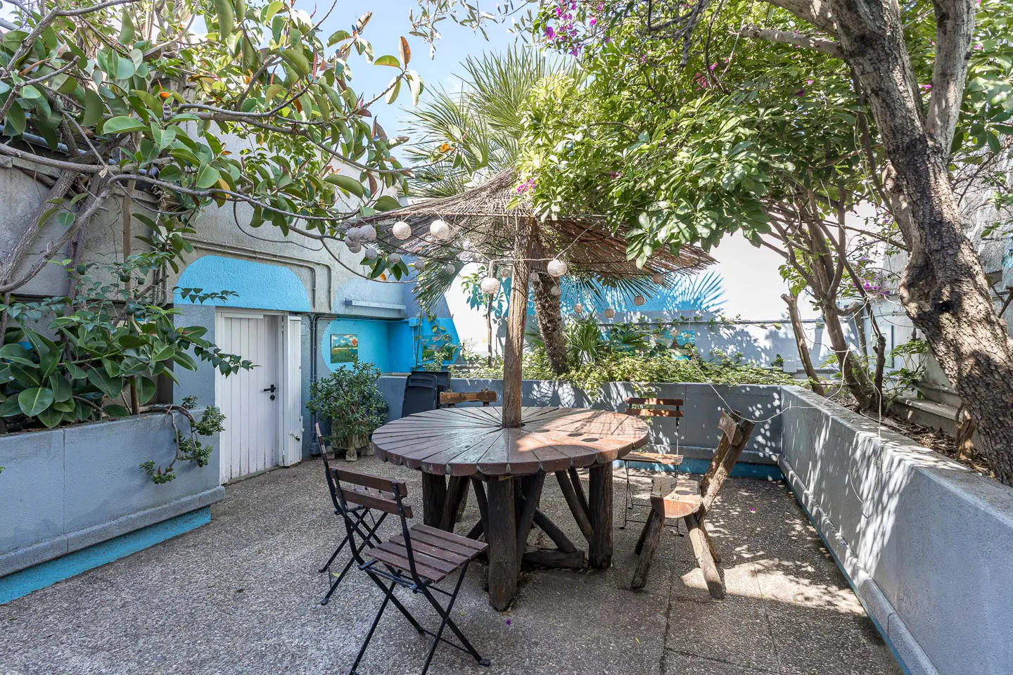 Outdoor patio with a round wooden table, chairs, and a straw umbrella. Green plants and trees surround the space. Blue accents on the walls.