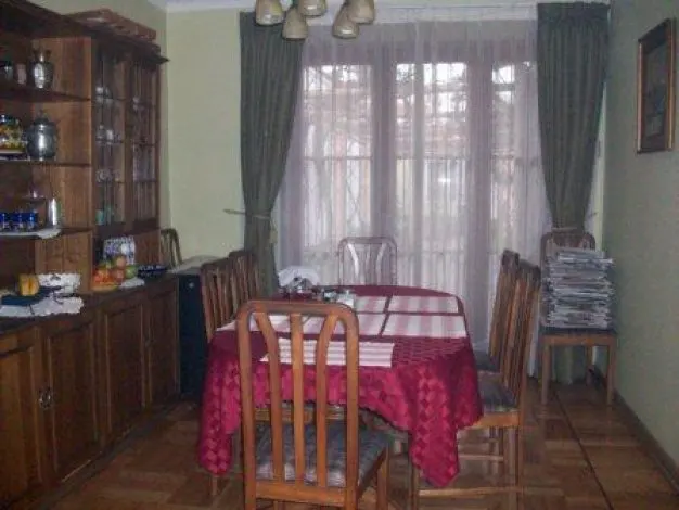 Dining room with wood floors, a red tablecloth on the table, and a wooden hutch filled with dishes. Green curtains frame the window.