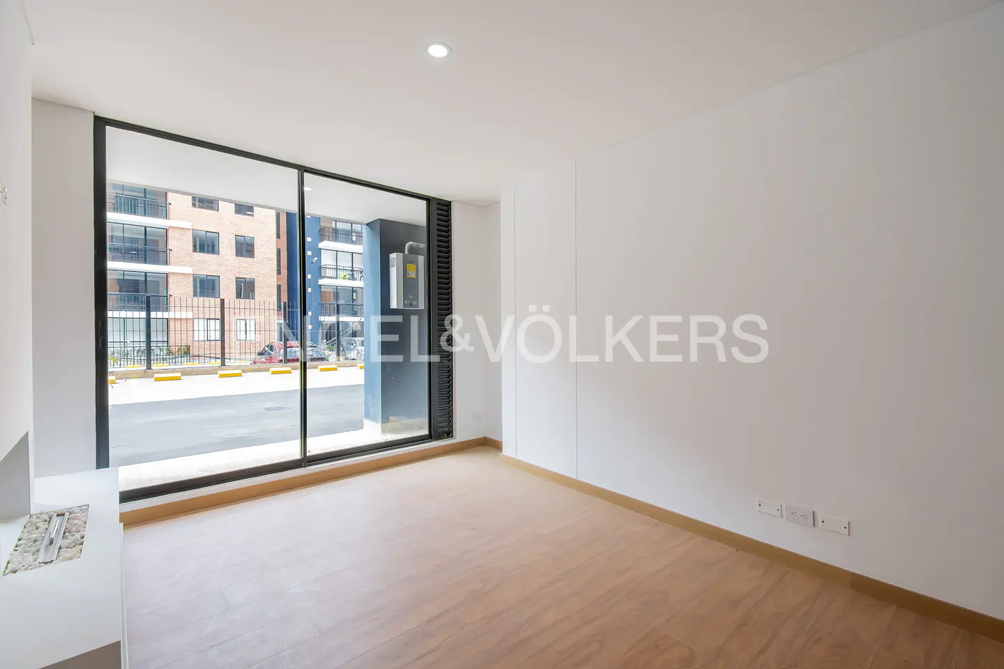 Empty room with light wood floors, white walls, and a large window. Outside is a brick building and parking area. Engel & Völkers logo on the wall.