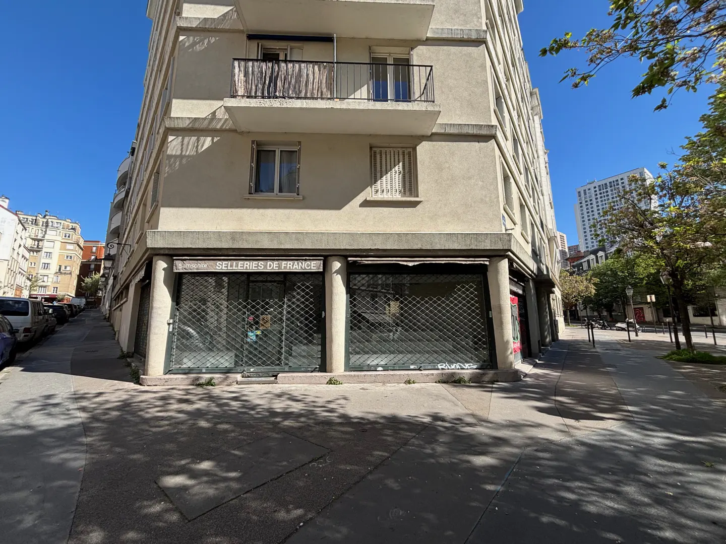 Exterior view of a beige apartment building with a storefront, "Selleries de France," secured by metal shutters on a sunny day.