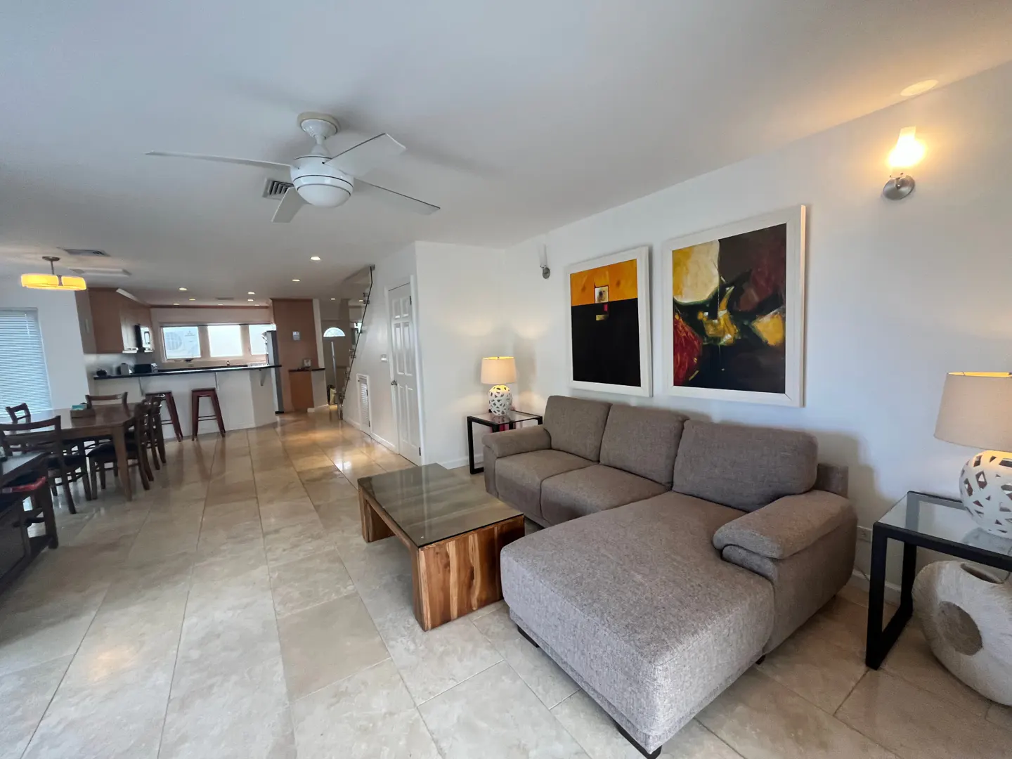 Open-concept living space with a gray sectional sofa, wood coffee table, and abstract art. Kitchen and dining area visible in the background.
