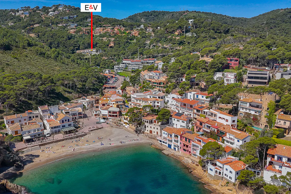 Aerial view of a coastal town with white buildings, red roofs, a sandy beach, and turquoise water. Hills covered in green trees surround the town.