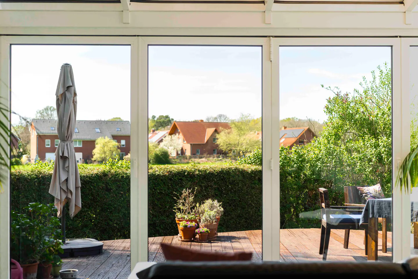 View through white framed glass doors to a wooden deck with potted plants, patio furniture, and a closed beige umbrella. Houses and greenery are visible beyond the deck.