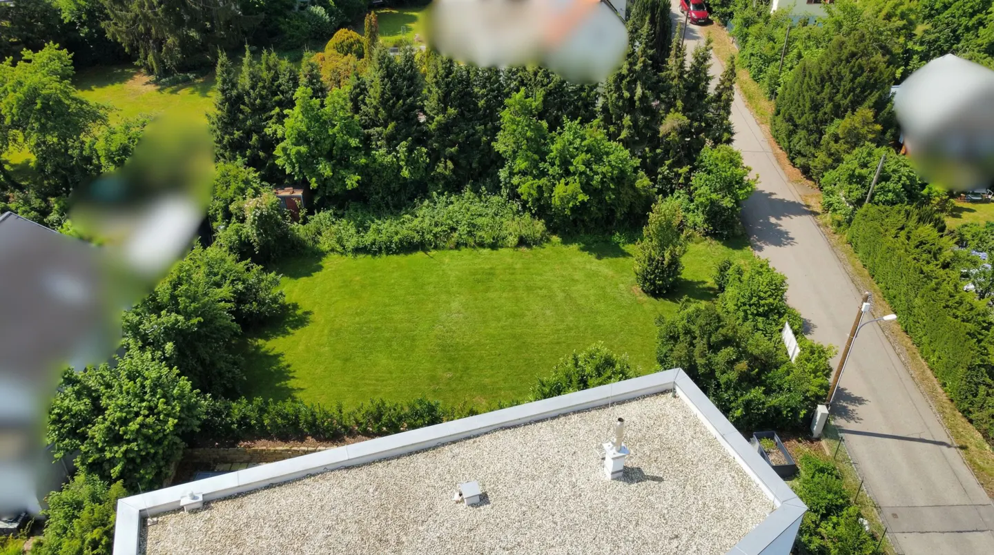Aerial view of a grassy lot surrounded by trees, next to a road. A building with a gravel roof is in the foreground.