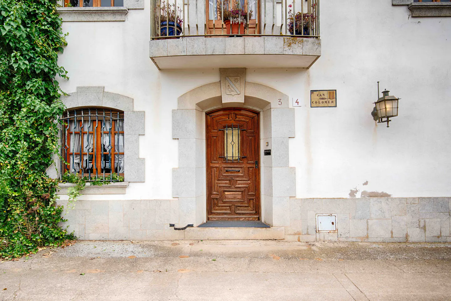 White stone building facade with a carved wooden door, ivy-covered wall, barred window, and a balcony with potted plants.
